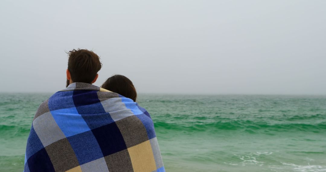 Caucasian Couple Wrapped in Blanket Facing Ocean on Overcast Beach
