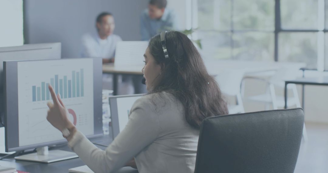 Female analyst presenting data on monitor, pointing and wearing headset in open office