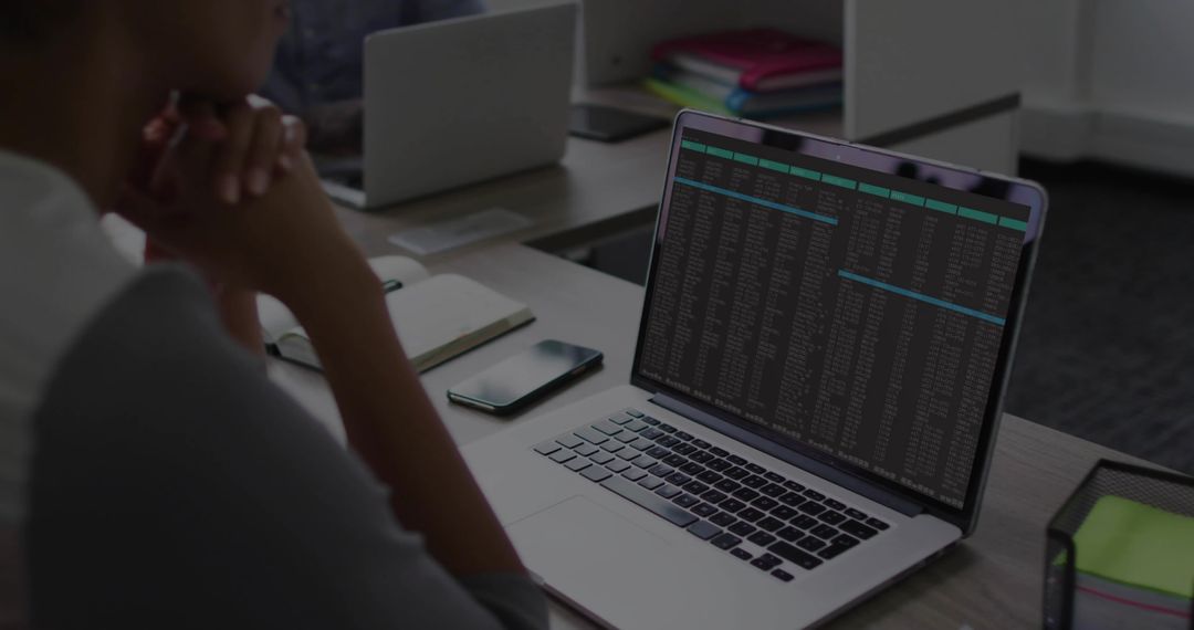 Developer Reviewing Terminal Output on Laptop in Modern Open Office with Notebook and Phone