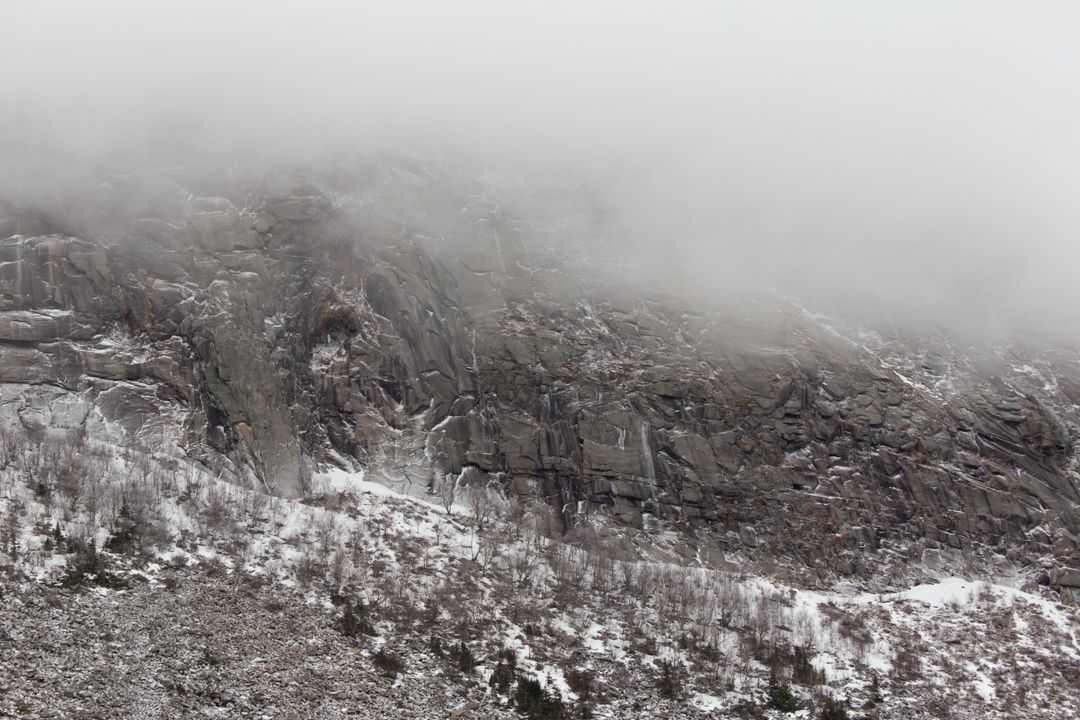 Fog-shrouding granite cliff looming over snow-dusted scree with sparse winter shrubs