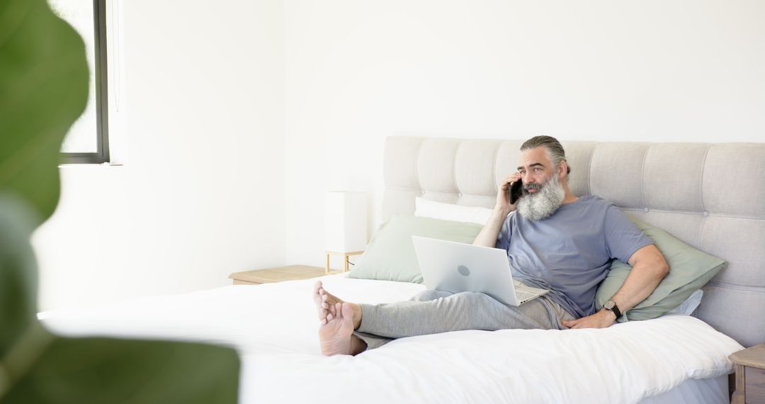 Senior Man Relaxing on Bed with Laptop and Smartphone in Modern Bright Bedroom