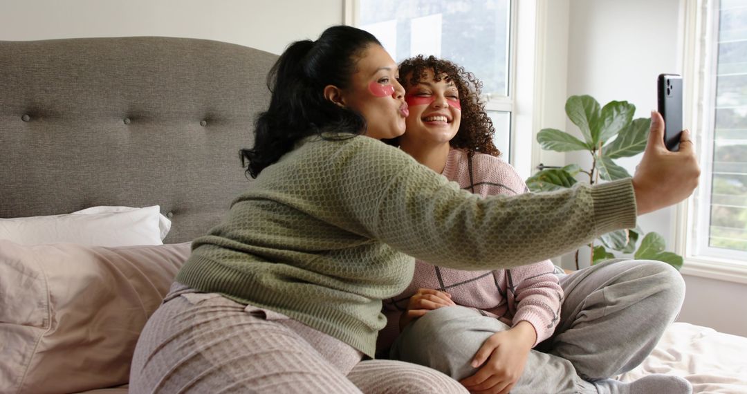 Friends taking selfie on bed with under-eye patches together enjoying cozy skincare moment