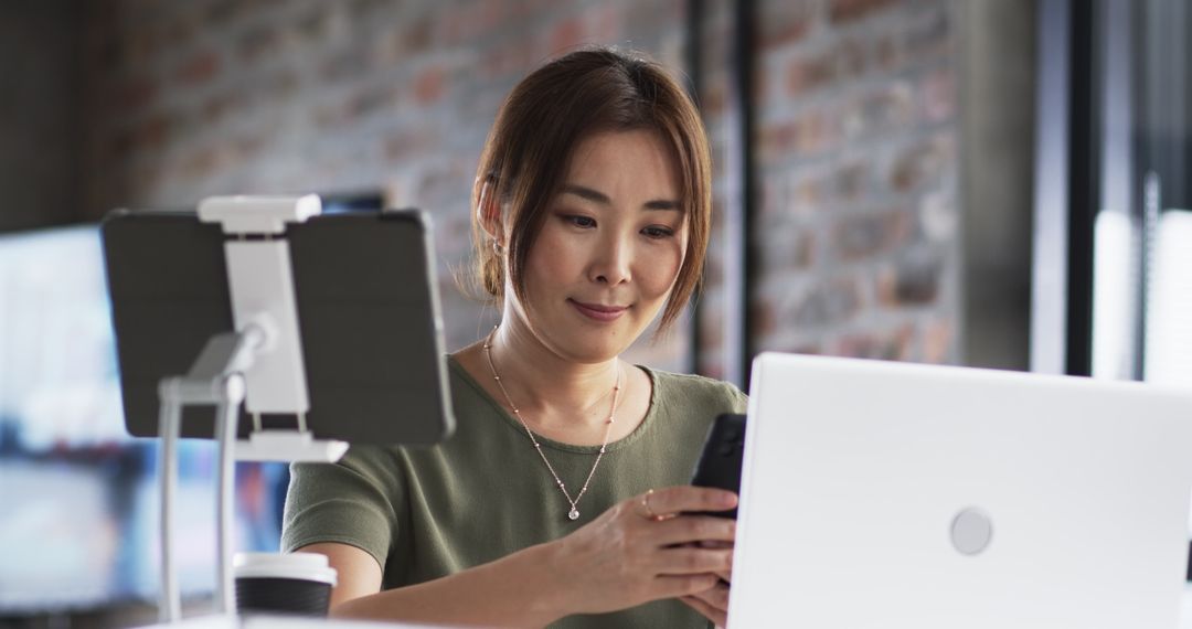 Middle-aged asian woman multitasking with laptop and smartphone in modern office