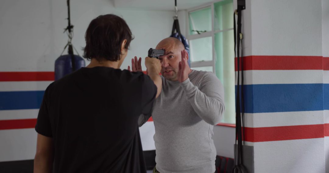 Man raising hands confronting partner pointing training pistol inside boxing gym