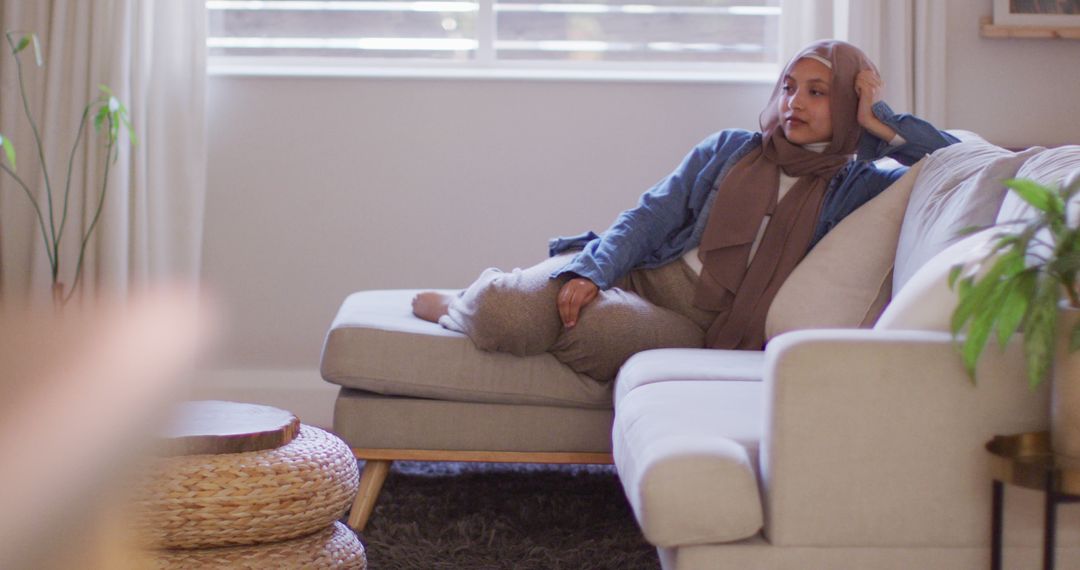 Image of smiling biracial woman in hijab relaxing on sofa at home - Free Images, Stock Photos and Pictures on Pikwizard.com