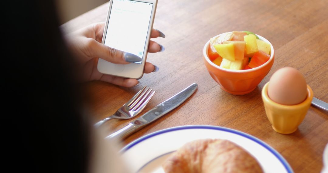 Hand using smartphone while having breakfast with croissant and fruit salad - Free Images, Stock Photos and Pictures on Pikwizard.com