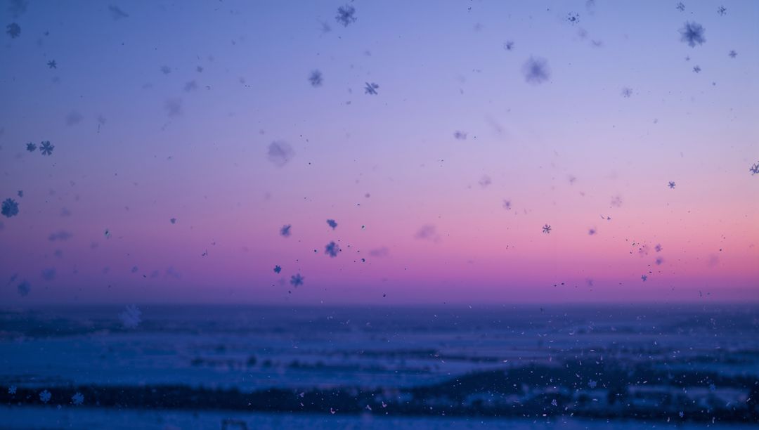 Frosted window with delicate snowflakes falling against pastel sunset over ocean horizon