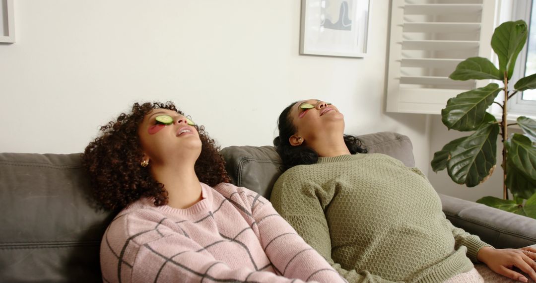 Female Friends Reclining on Sofa with Cucumber Eye Slices Relaxing Self-Care at Home