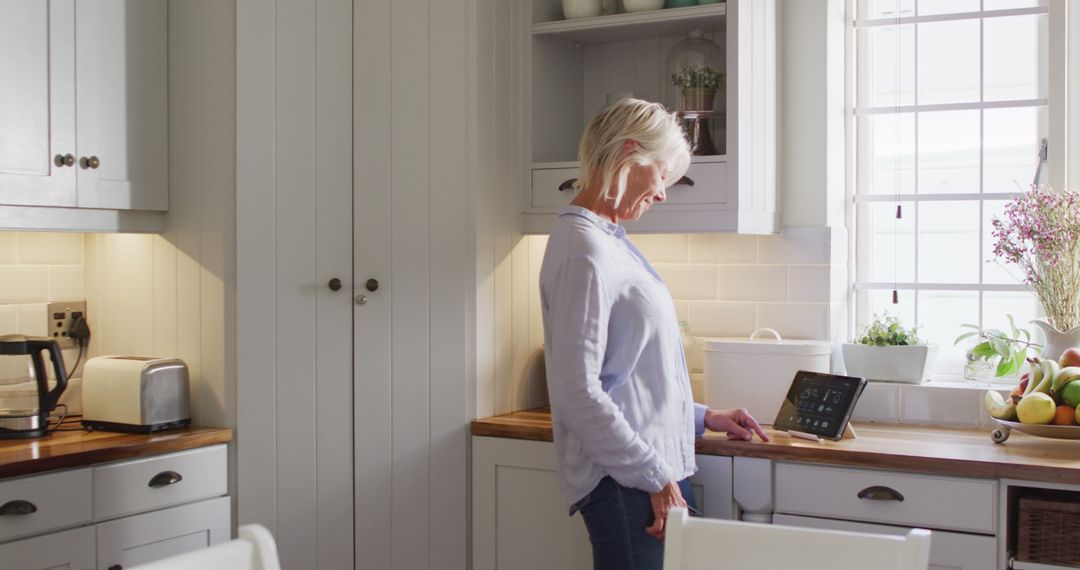 Happy senior caucasian woman standing in kitchen, using smartphone and tablet - Free Images, Stock Photos and Pictures on Pikwizard.com