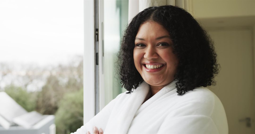 Smiling woman in white bathrobe relaxing by open sliding door enjoying morning light