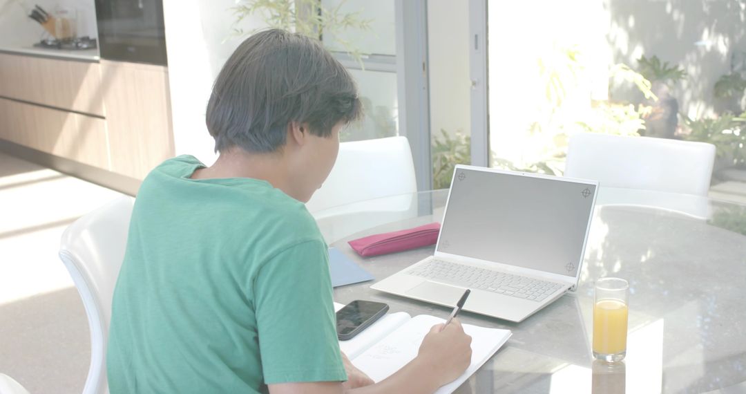 Asian man writing and studying at home dining table with laptop, smartphone and orange juice