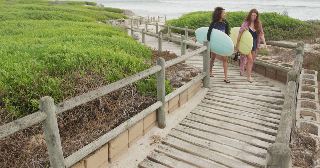 Two Women Walking with Surfboards on Wooden Path near Ocean - Free Images, Stock Photos and Pictures on Pikwizard.com