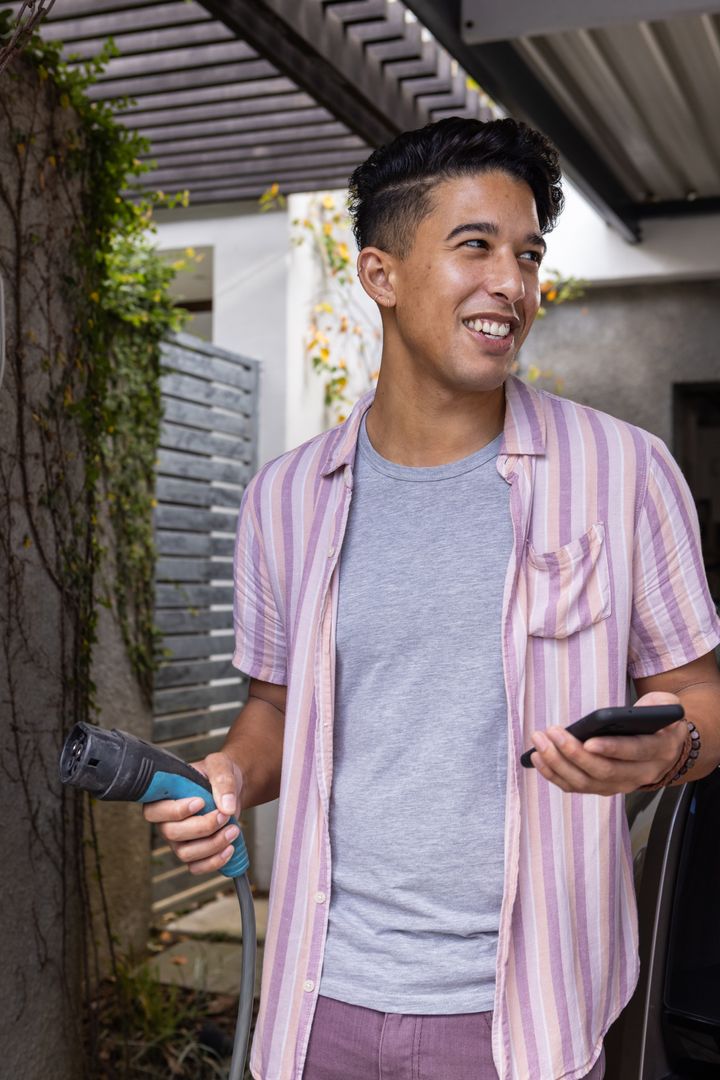 Young Man Charging Electric Vehicle and Using Smartphone in Carport
