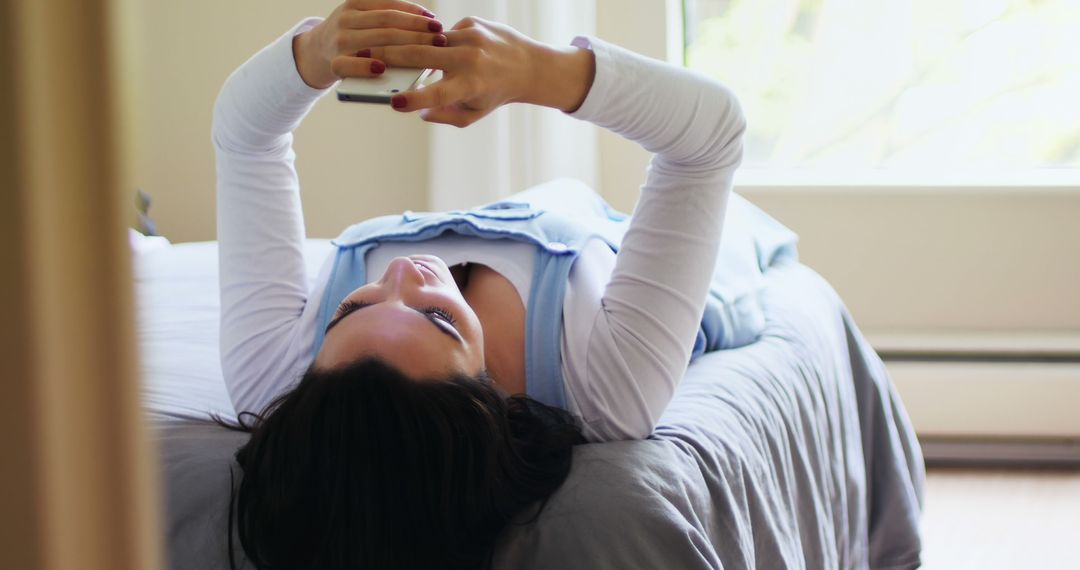 Young woman lying on bed holding smartphone in relaxed setting - Free Images, Stock Photos and Pictures on Pikwizard.com