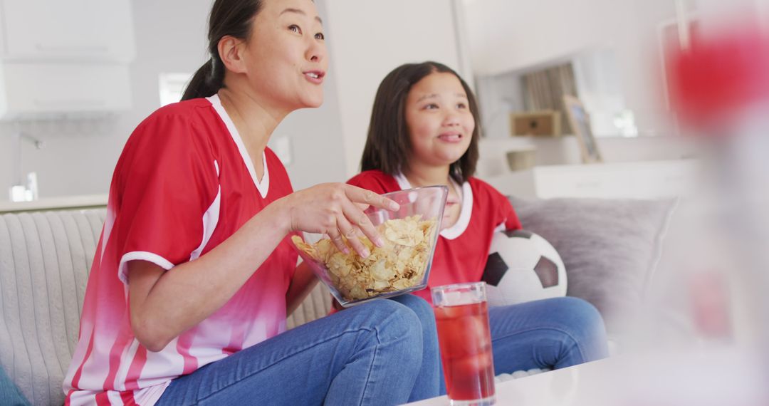 Image of happy asian mother and daughter eating chips and watching match in tv - Free Images, Stock Photos and Pictures on Pikwizard.com