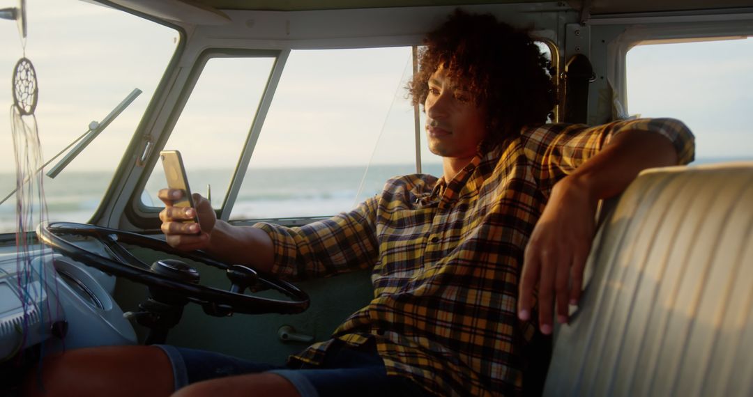 Young man relaxing in van using smartphone near beach during sunset - Free Images, Stock Photos and Pictures on Pikwizard.com