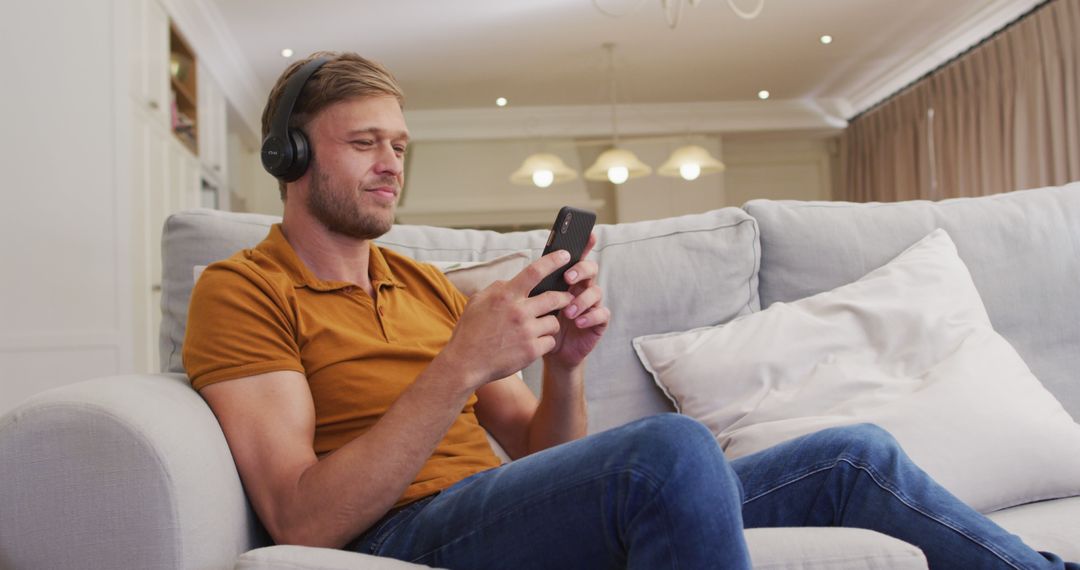 Portrait of happy caucasian man sitting on sofa wearing headphones and using smartphone - Free Images, Stock Photos and Pictures on Pikwizard.com