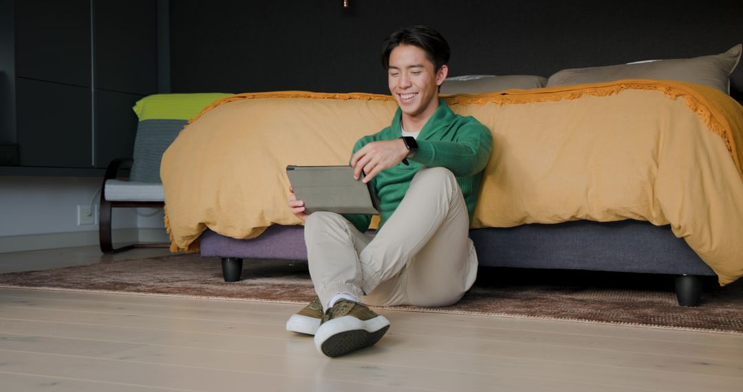Modern Asian Man Relaxing in Bedroom with Tablet and Smartwatch