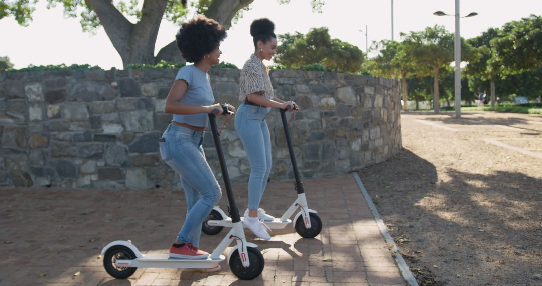 Two Young Women Riding Electric Scooters in Park on Sunny Day - Free Images, Stock Photos and Pictures on Pikwizard.com