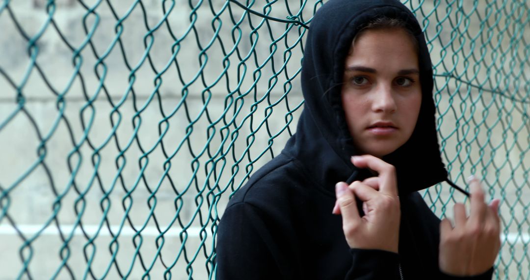 A young Caucasian girl wearing a black hoodie stands behind a chain-link fence, with copy space - Free Images, Stock Photos and Pictures on Pikwizard.com
