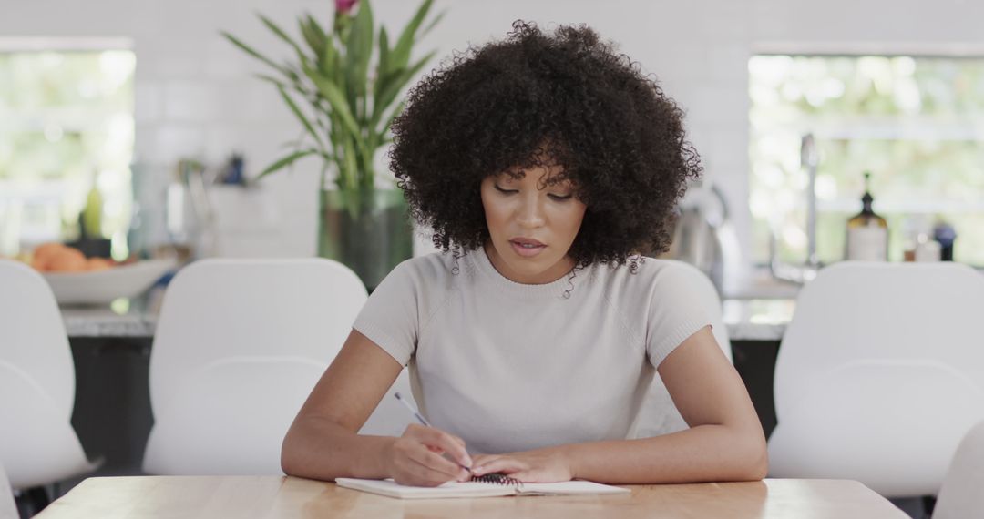 Woman with Curly Hair Writing on Notebook at Kitchen Table - Free Images, Stock Photos and Pictures on Pikwizard.com