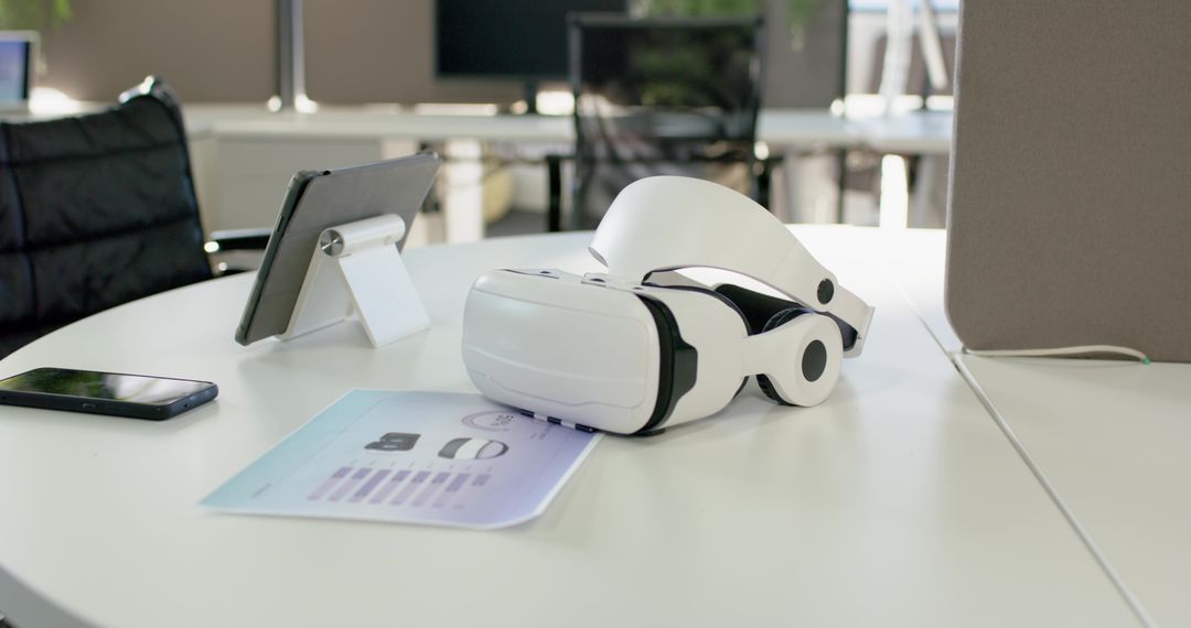 White VR headset resting on spec sheet on round meeting table with tablet and phone in open office