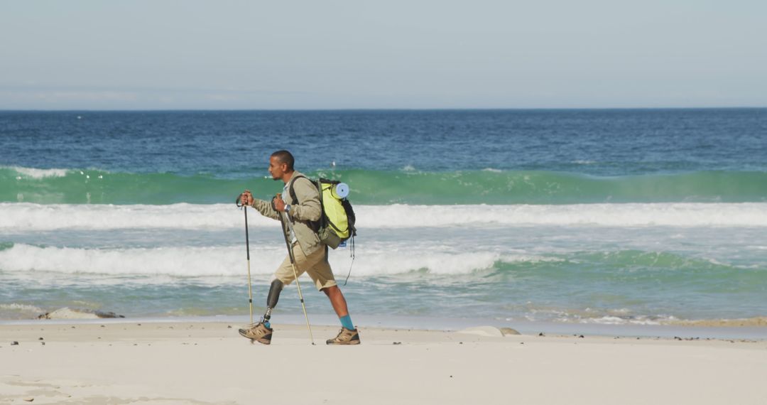 Biracial man with prosthetic leg trekking with backpack and walking poles on a beach by the sea - Free Images, Stock Photos and Pictures on Pikwizard.com