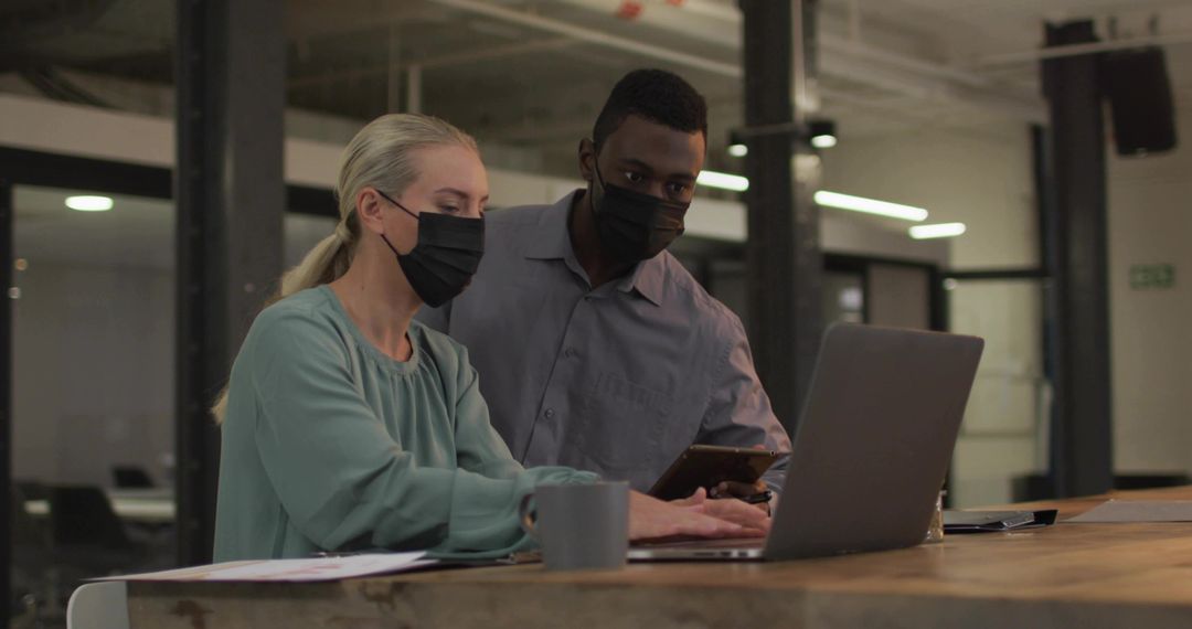 Masked coworkers collaborating over laptop and smartphone in modern open-plan office team