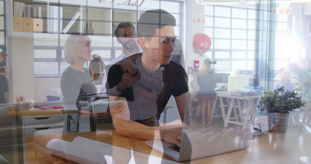Asian Man Multitasking on Laptop Amidst Busy Office Environment