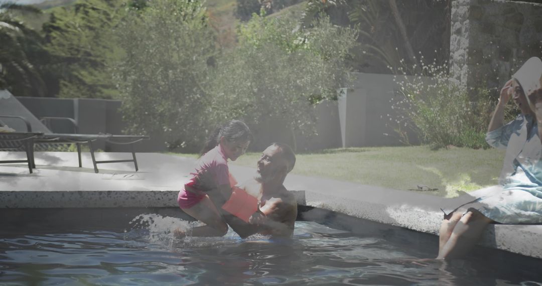 Smiling father lifting daughter in backyard pool, splashing during sunny summer day