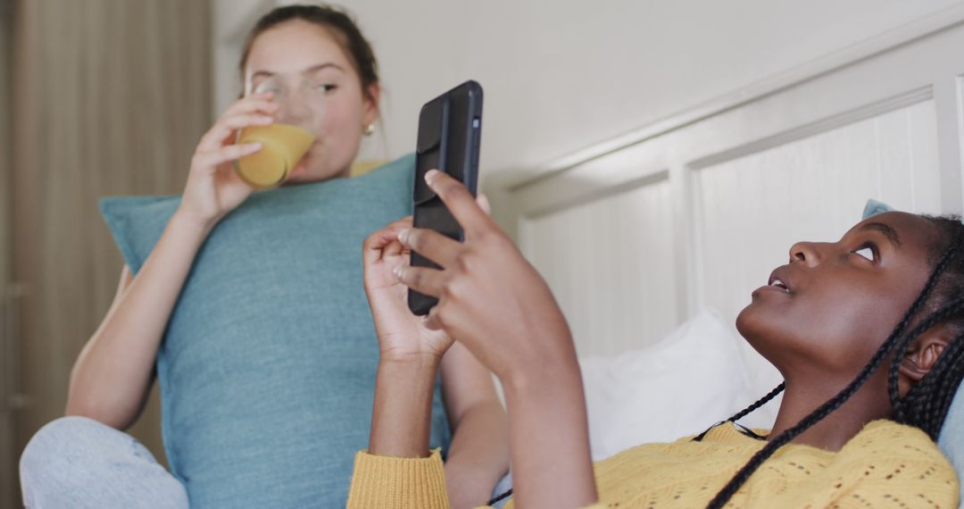 Two Young Women Relaxing on Bed Using Smartphone and Drinking Juice - Free Images, Stock Photos and Pictures on Pikwizard.com