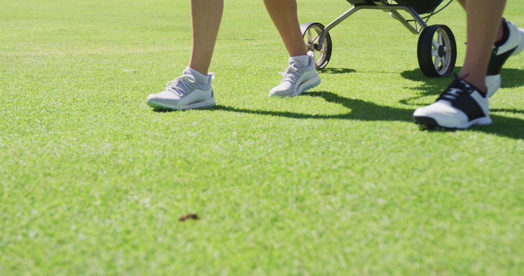 Two People Walking Across Green Grass Pushing Golf Cart on Sunny Day - Free Images, Stock Photos and Pictures on Pikwizard.com