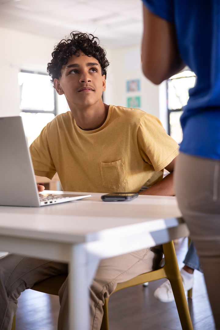 Teenage Boy in Classroom Using Laptop with Teacher Present