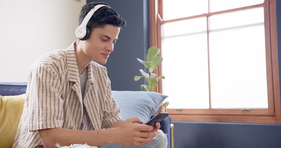 Young Man Relaxing with Headphones Using Smartphone near Window