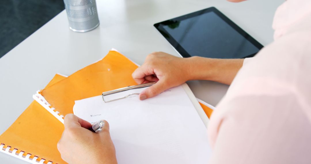 Person Filling Out Form on Clipboard with Tablet Nearby - Free Images, Stock Photos and Pictures on Pikwizard.com