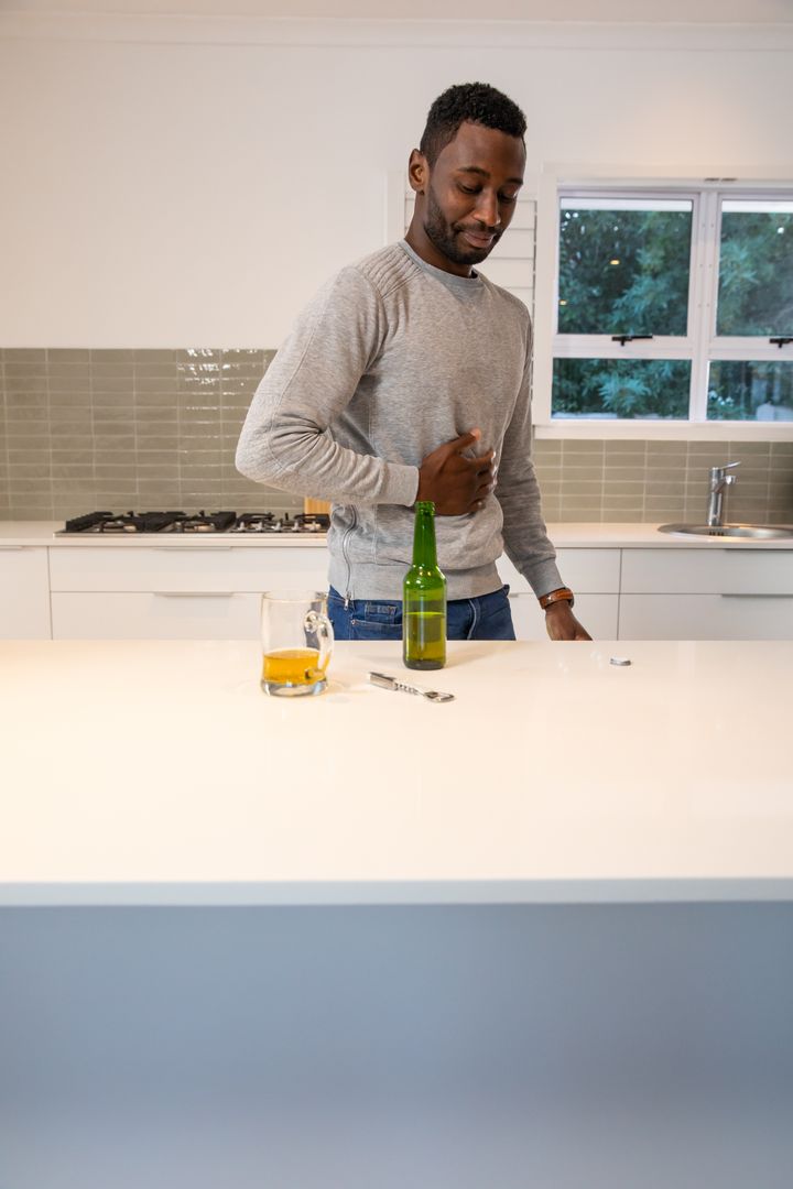 Man Feeling Discomfort Holding Stomach Near Beer in Modern Kitchen