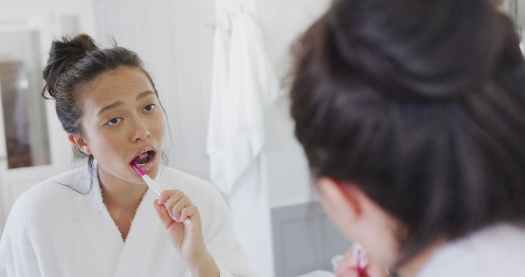 Woman brushing teeth in bathroom mirror wearing white robe - Free Images, Stock Photos and Pictures on Pikwizard.com