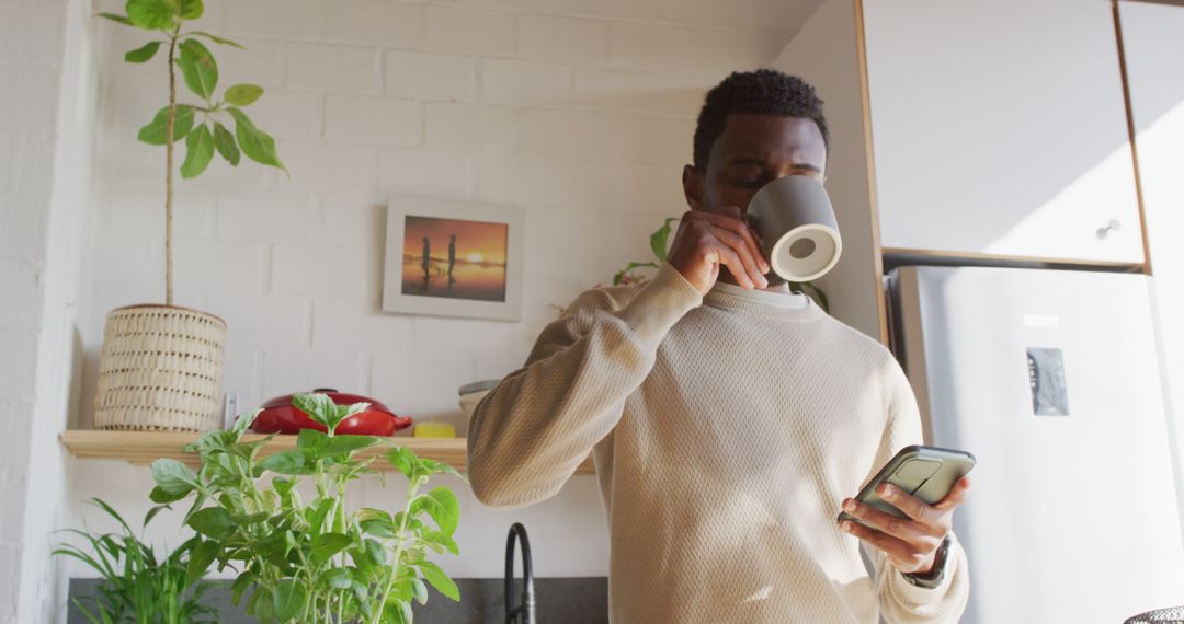 Happy african american man standing in kitchen, drinking coffee and using smartphone - Free Images, Stock Photos and Pictures on Pikwizard.com