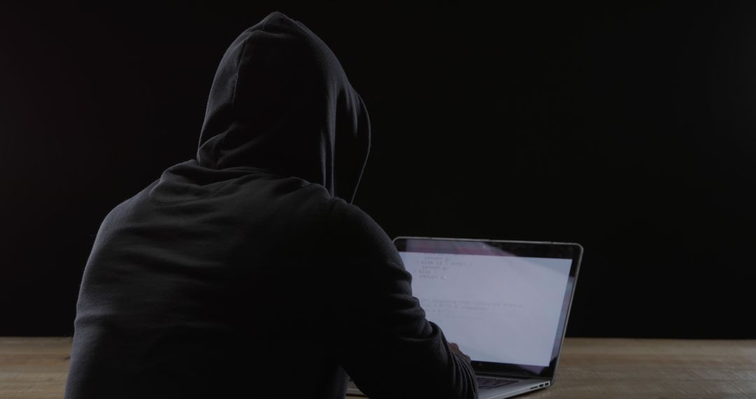 Hooded figure works on a laptop in a dimly lit room - Free Images, Stock Photos and Pictures on Pikwizard.com
