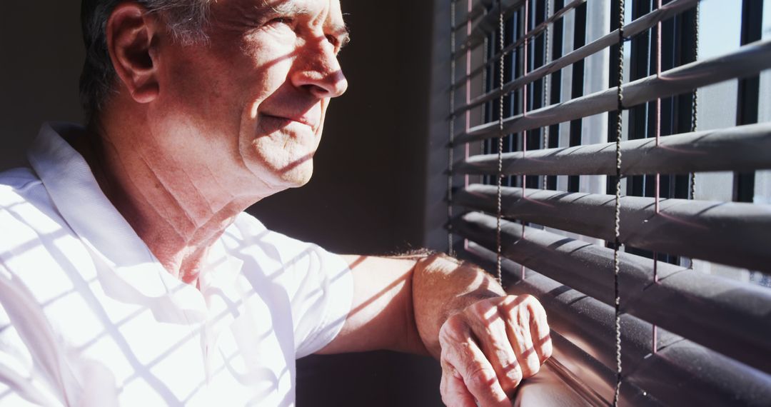 Pensive Senior Man Looking Out Window Through Blinds in Sunlit Home - Free Images, Stock Photos and Pictures on Pikwizard.com