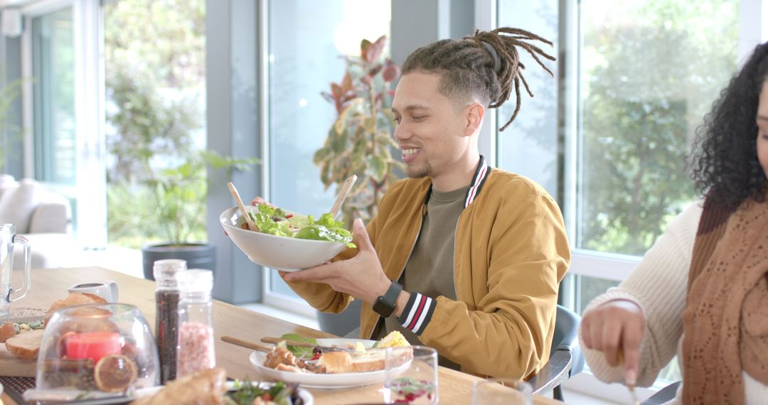 Diverse friends enjoying casual brunch around sunlit table, man serving fresh salad bowl