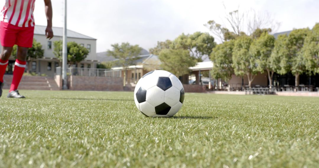 Close-up rolling soccer ball on trimmed grass with player approaching in red-striped jersey