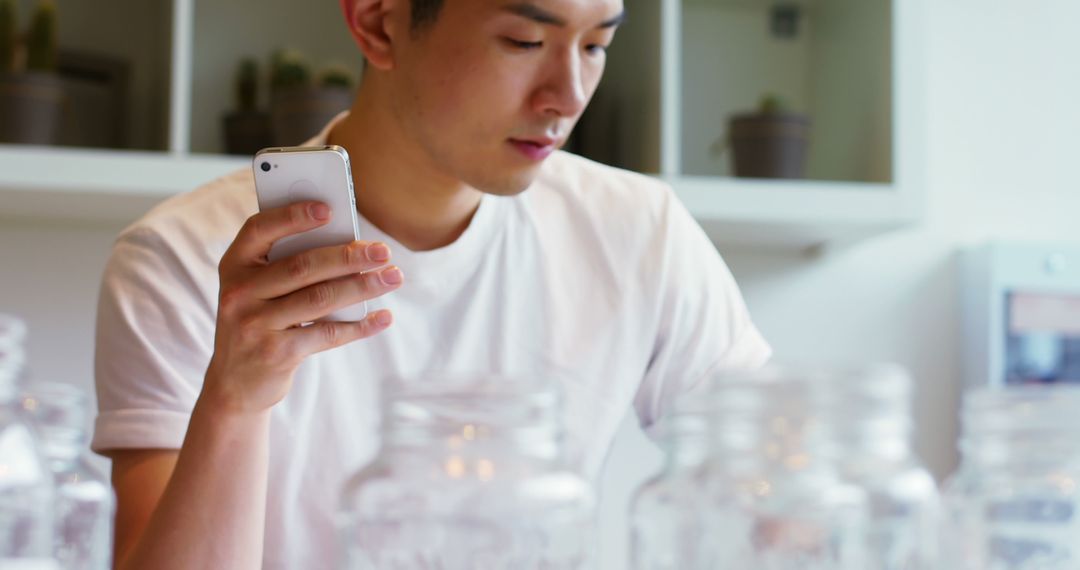 Young Man in White T-shirt Focusing on Smartphone amid Glass Jars - Free Images, Stock Photos and Pictures on Pikwizard.com