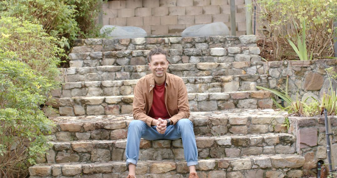 Smiling man sitting on rustic stone steps in backyard garden wearing brown jacket