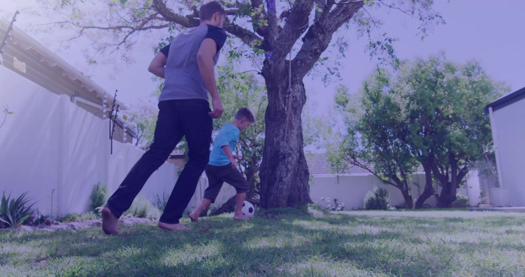 Father and son playing soccer barefoot on backyard lawn under shady tree