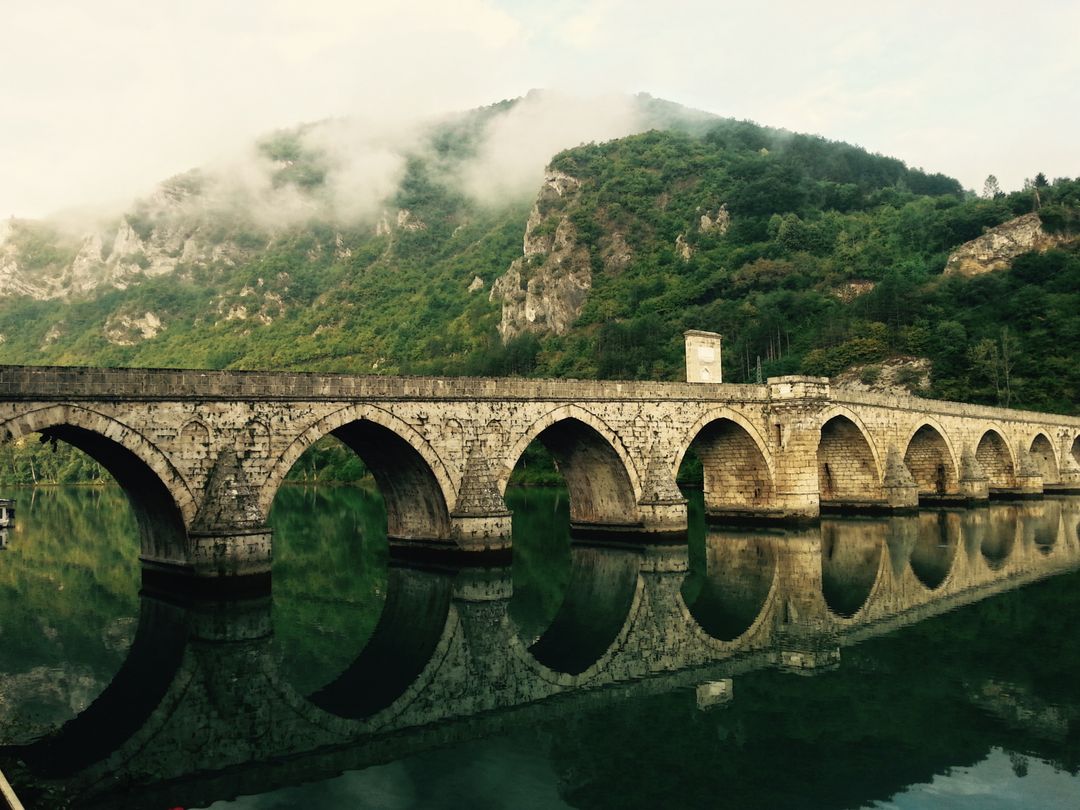 Historical Stone Bridge Reflected in Calm River with Misty Mountains in Background - Free Images, Stock Photos and Pictures on Pikwizard.com