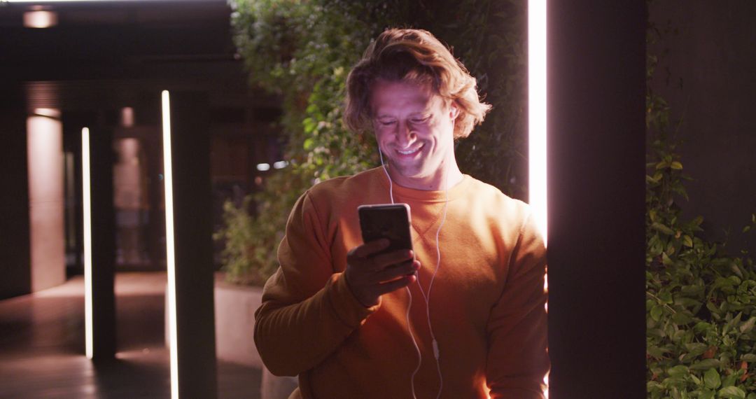 Smiling Man Using Smartphone with Earphones at Night in Urban Setting - Free Images, Stock Photos and Pictures on Pikwizard.com