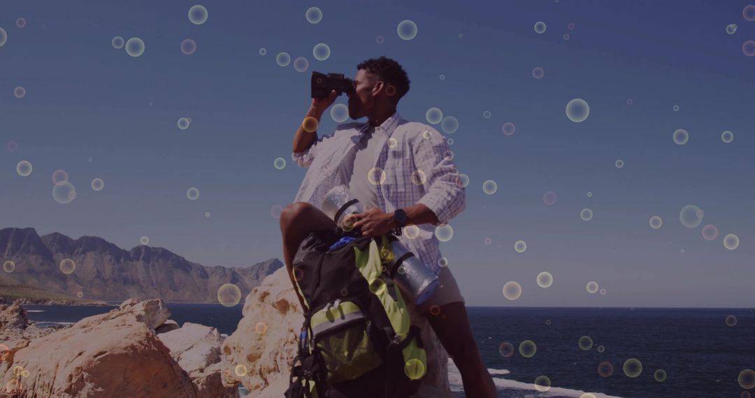 Scanning coastline with binoculars standing on rocky shore with backpack and thermos