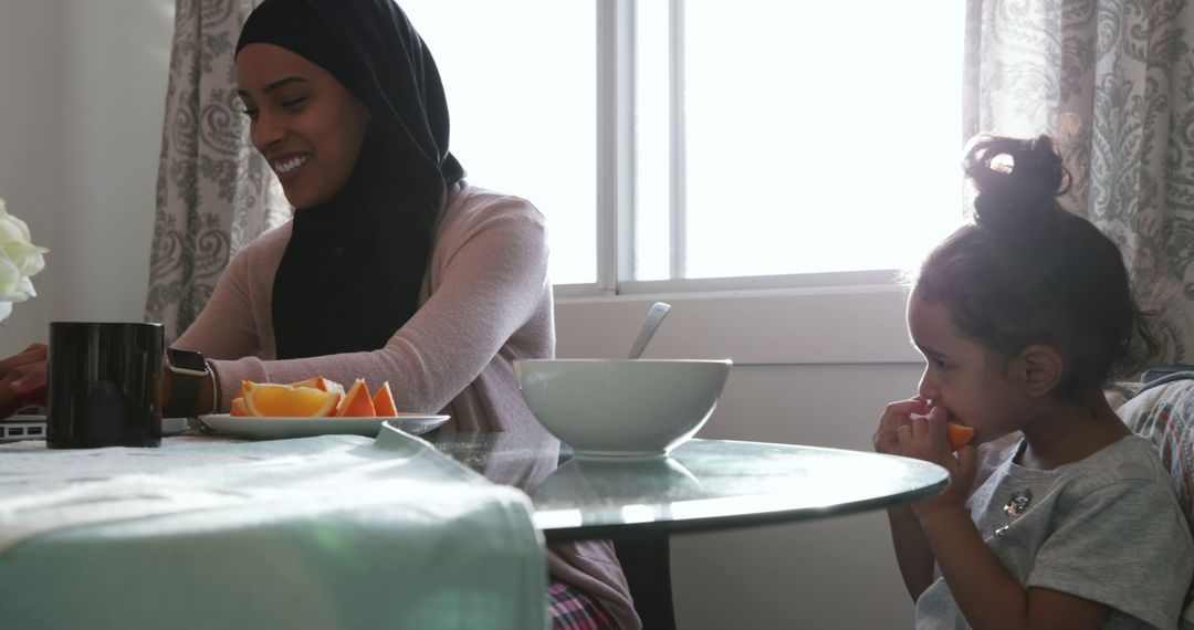 Mother in Hijab Using Laptop While Daughter Eats Orange at Home