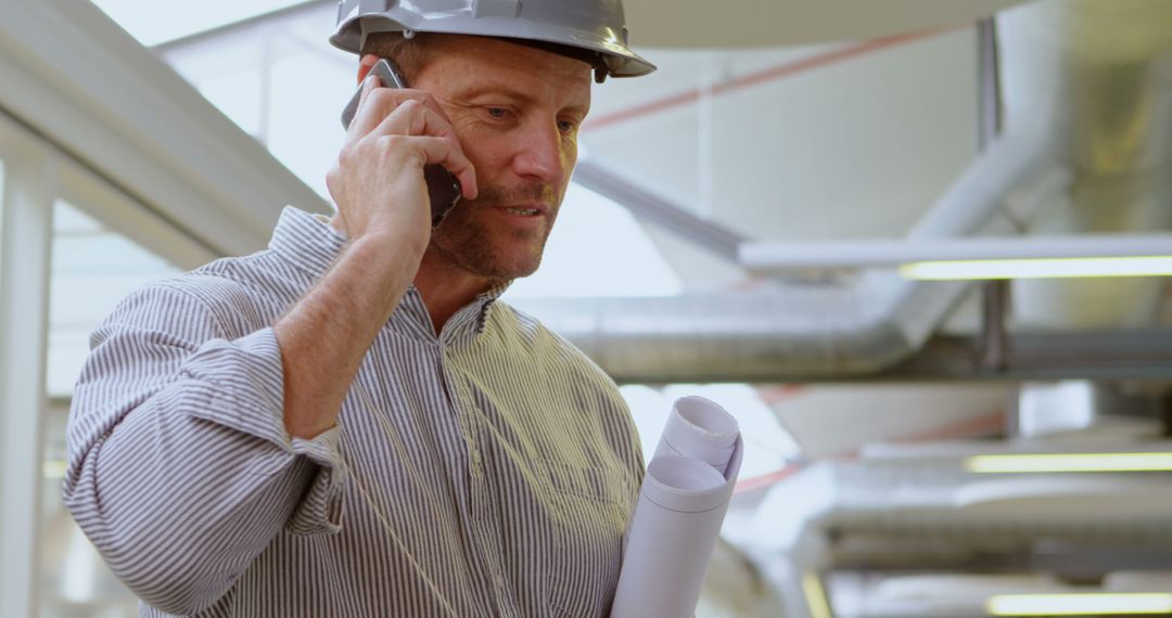 Engineer Discussing Project on Phone While Holding Blueprints in Modern Workspace - Free Images, Stock Photos and Pictures on Pikwizard.com